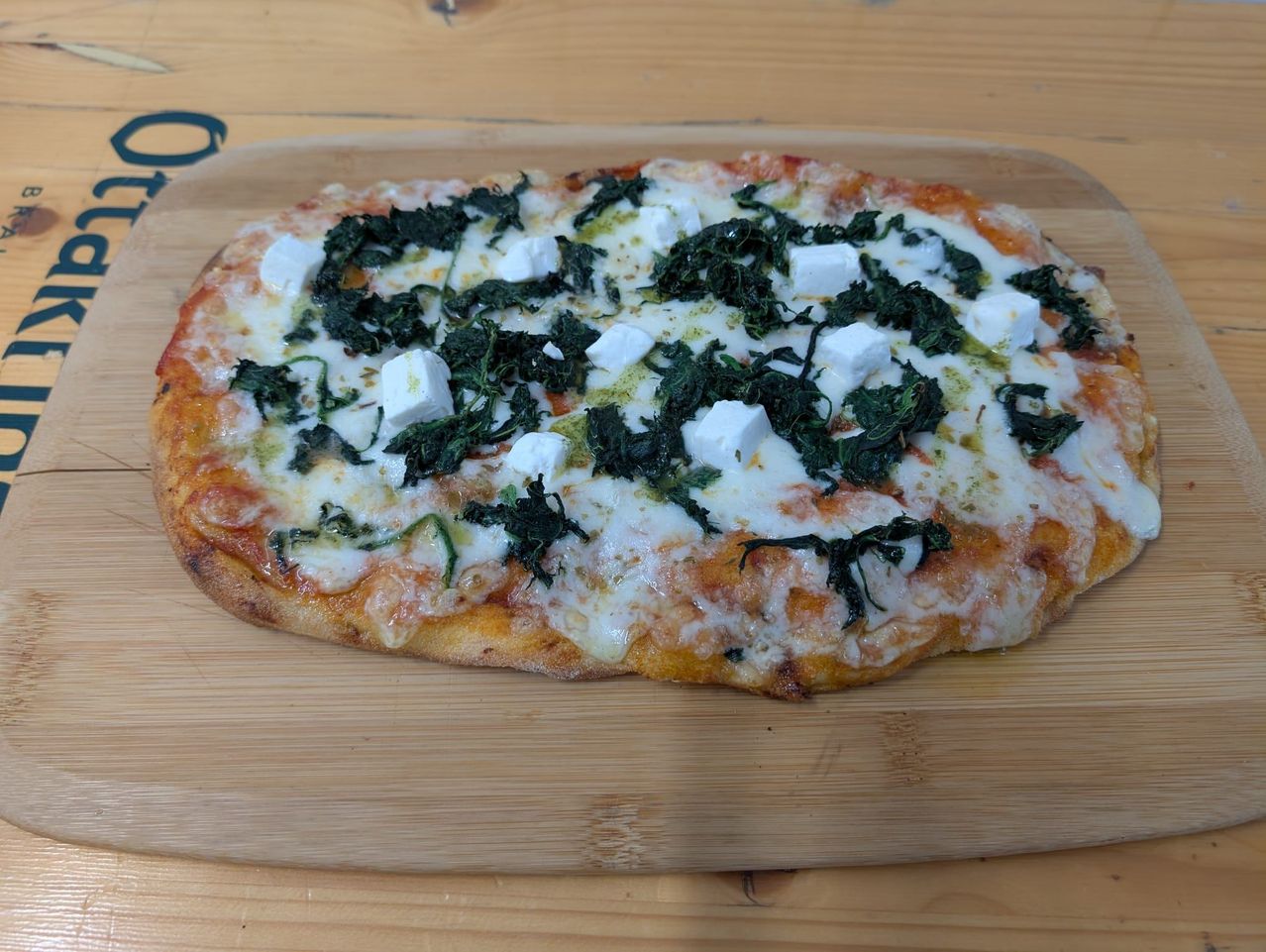 Eine Pizza mit geschmolzenem Käse, Spinat und Feta-Würfeln liegt auf einem hölzernen Schneidebrett.