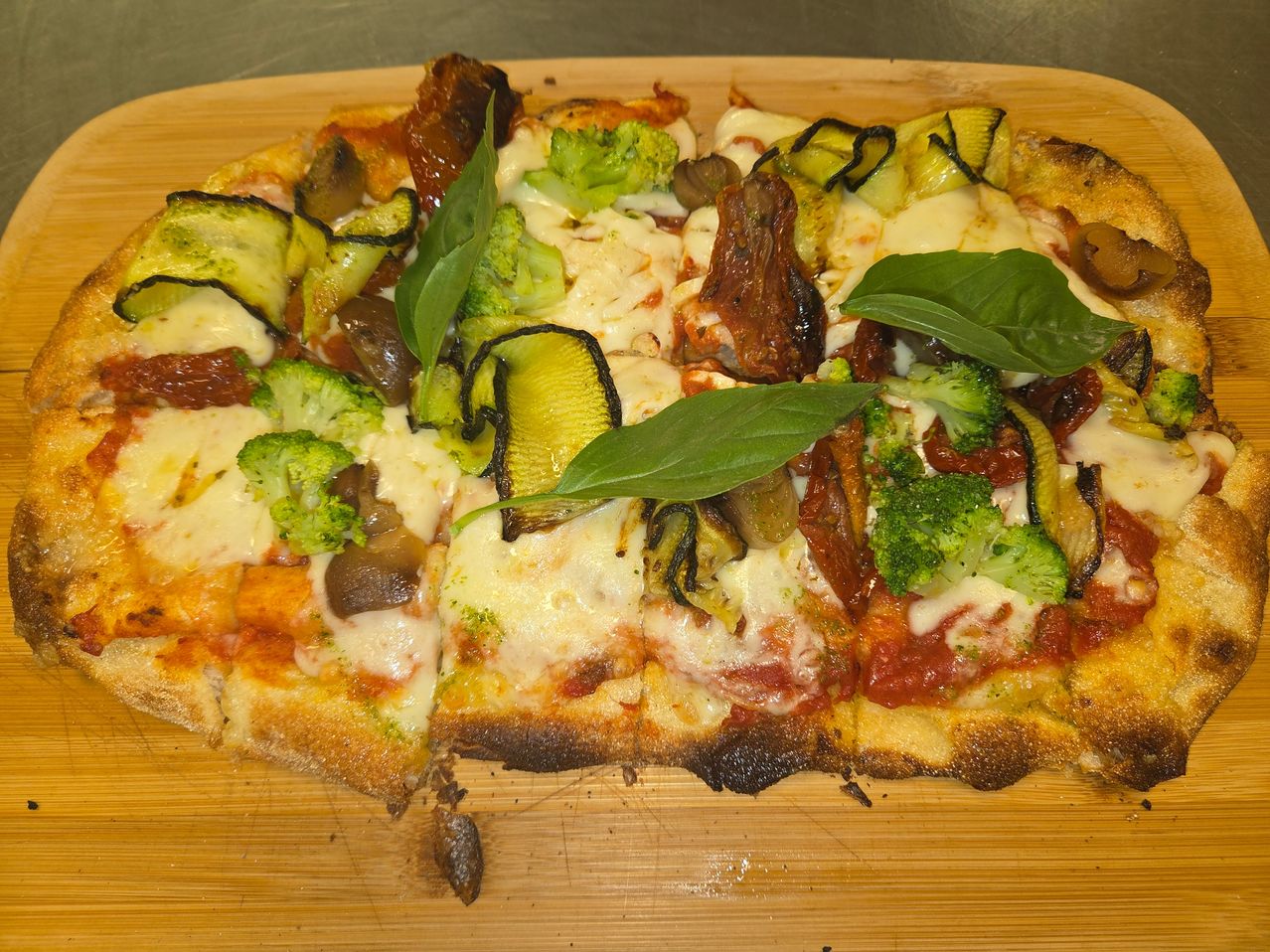 A close-up of a pizza with various toppings including broccoli, mushrooms, and basil leaves on a wooden chopping board.