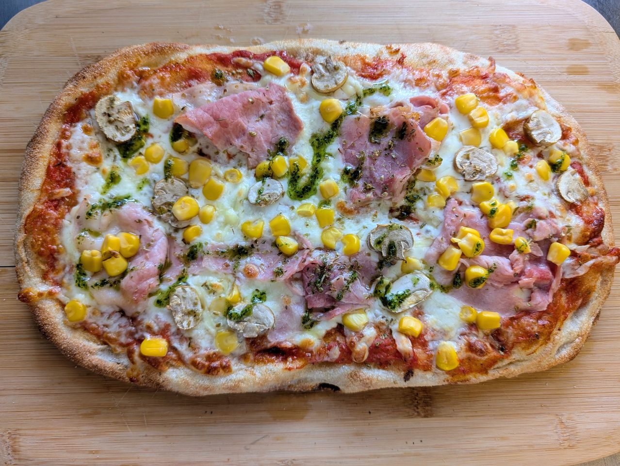 A round pizza topped with melted cheese, corn, mushrooms, and ham slices. It is placed on a wooden surface.