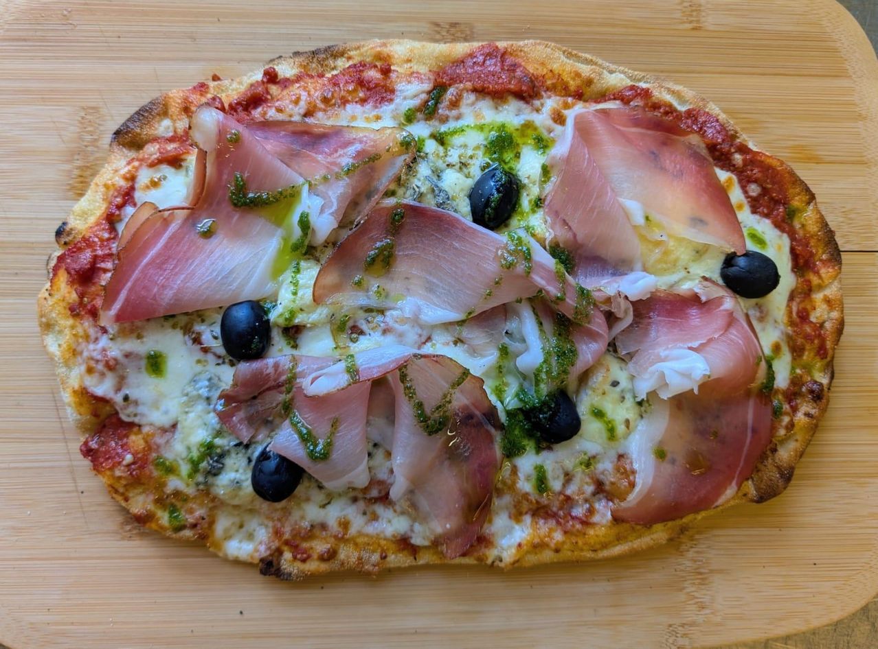 Eine ganze Pizza mit Käse, Oliven und Prosciutto. Eine grüne Sauce ist über die Pizza verteilt.