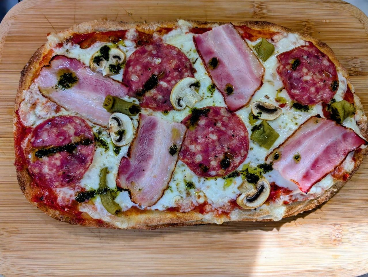 Eine rechteckige Pizza mit Schinken, Salami, Champignons und grünen Paprikaschoten auf einer Holzunterlage.