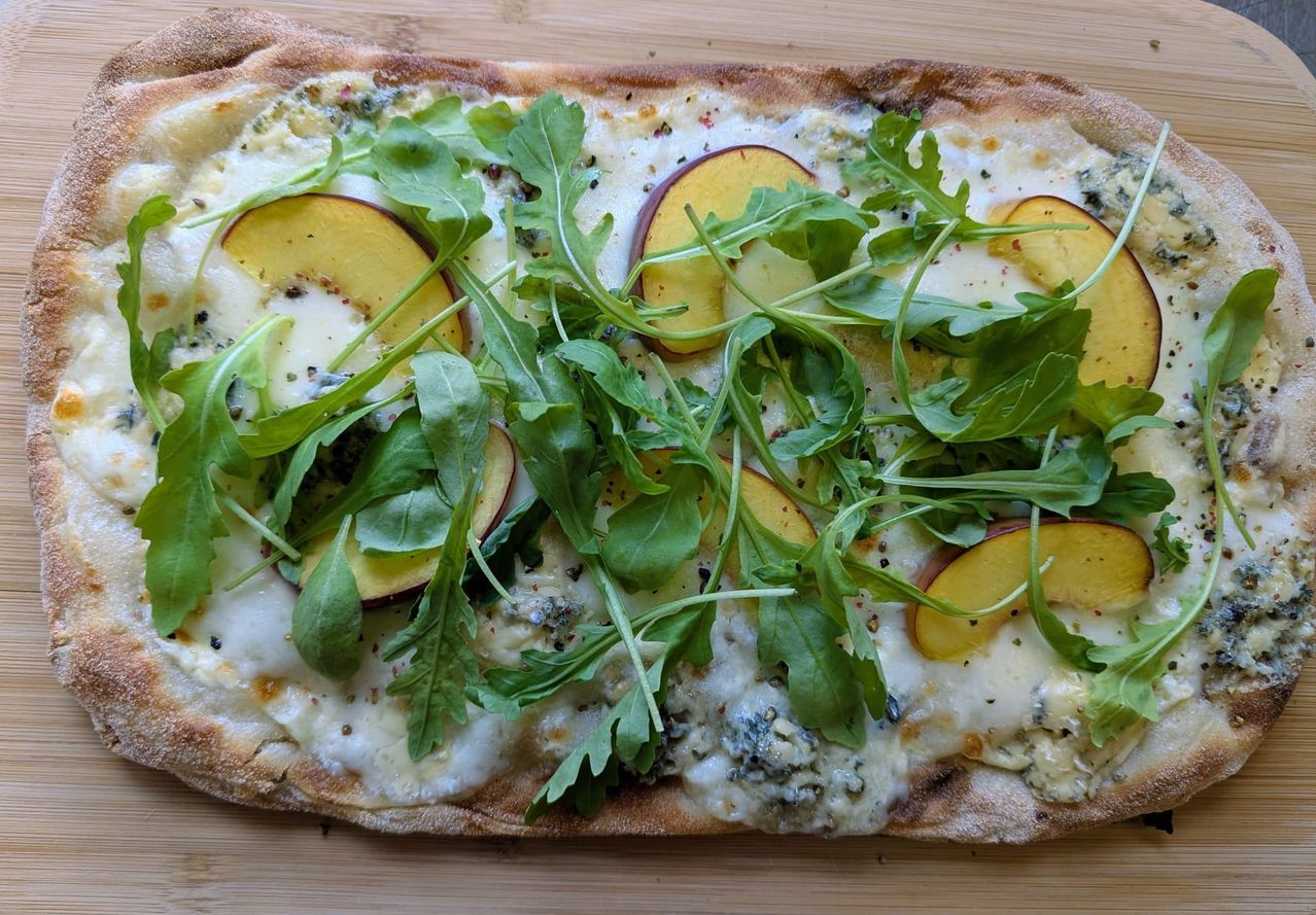 A flatbread pizza topped with slices of peach, blue cheese, and arugula on a wooden surface.