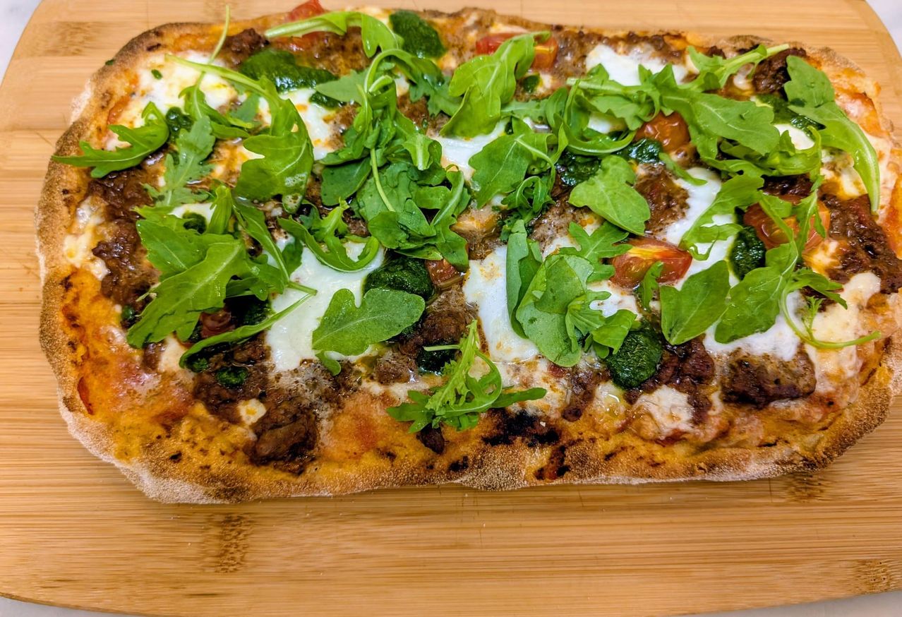 A close-up view of a pizza topped with melted cheese, ground beef, spinach, and arugula on a wooden board.