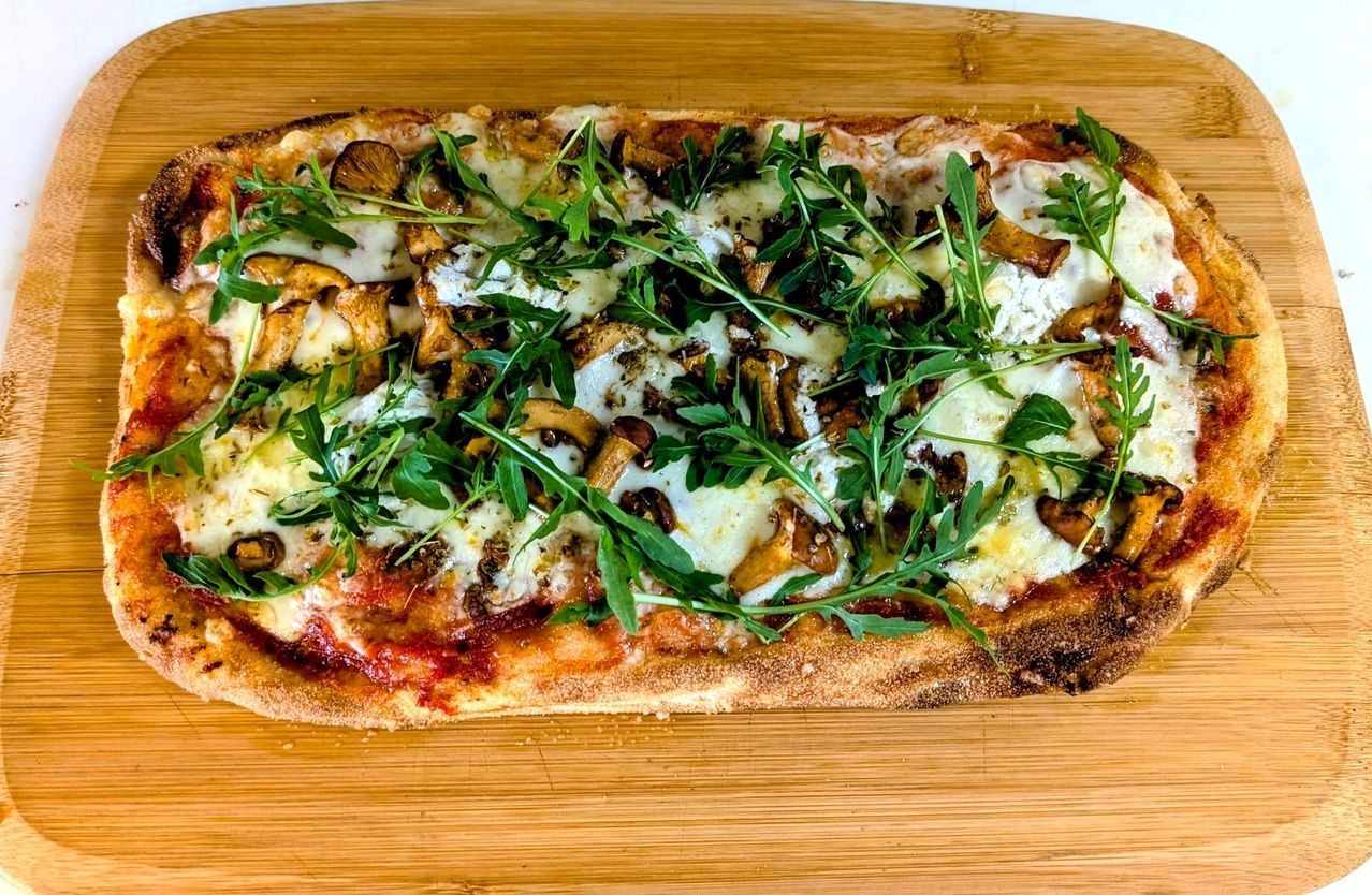 Eine Pizza mit Käse, Pilzen und Rucola auf einem Holzbrett, leicht angebrannt an den Rändern.