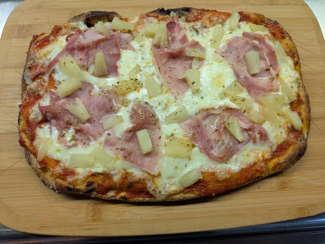 A pizza with melted cheese, ham slices, and pineapple chunks on a wooden chopping board.