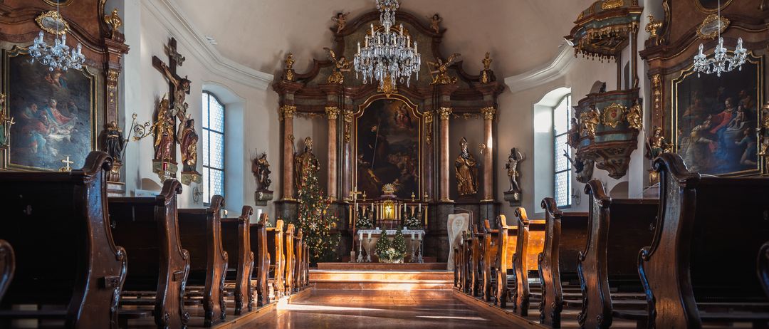 Ein leerer Kircheninnenraum mit Holzbänken, einem großen Altar und einer großen Kristallleuchte. Dekorationen und Statuen schmücken die Wände.