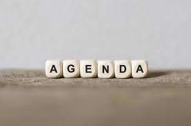 Das Wort Agenda in weißen Würfeln auf einer braunen Oberfläche geschrieben. Der Hintergrund ist schlicht und grau.