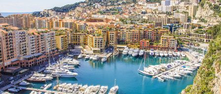 Luftaufnahme des Hafens von Monaco mit vielen angedockten Booten, farbenfrohen Gebäuden und einem Berg im Hintergrund.