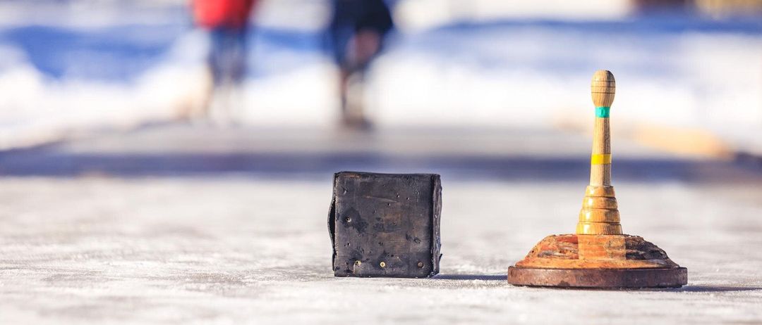 Ein Nahaufnahme eines hölzernen Objekts, das einem Kegel ähnelt, und einer kleinen, abgenutzten Box liegt auf einer verschneiten Oberfläche mit verschwommenen Figuren von Menschen im Hintergrund.