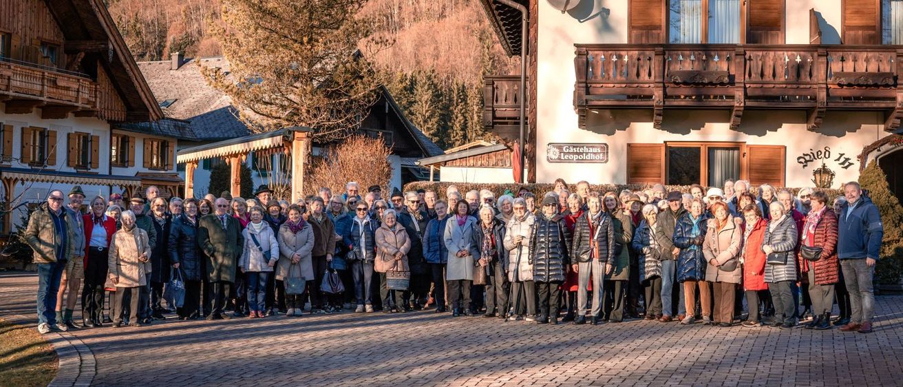 Eine Gruppe älterer Menschen steht vor einem Gebäude mit Balkonen. Dahinter befinden sich weitere Häuser, ein Baum und Berge.