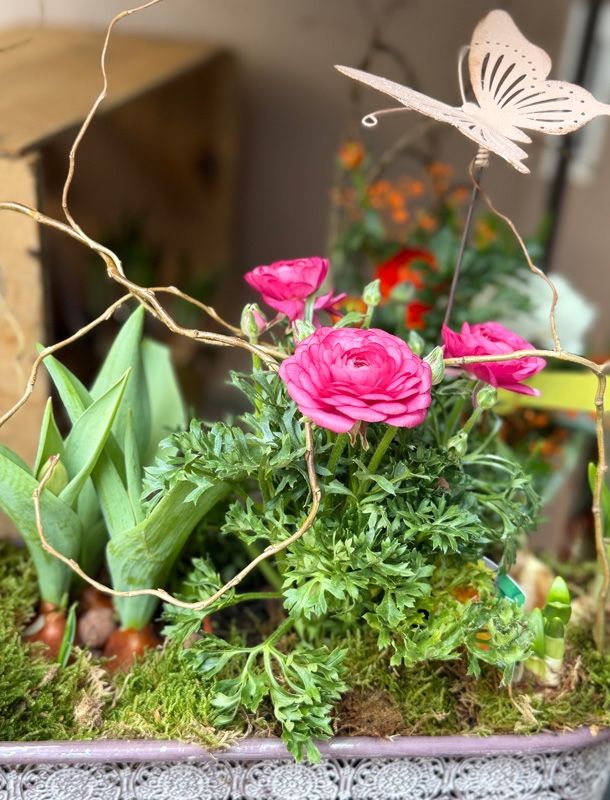 A close-up of a floral arrangement with pink roses, green leaves, and moss, with a butterfly on a stick. Other flowers in the background are blurred.
