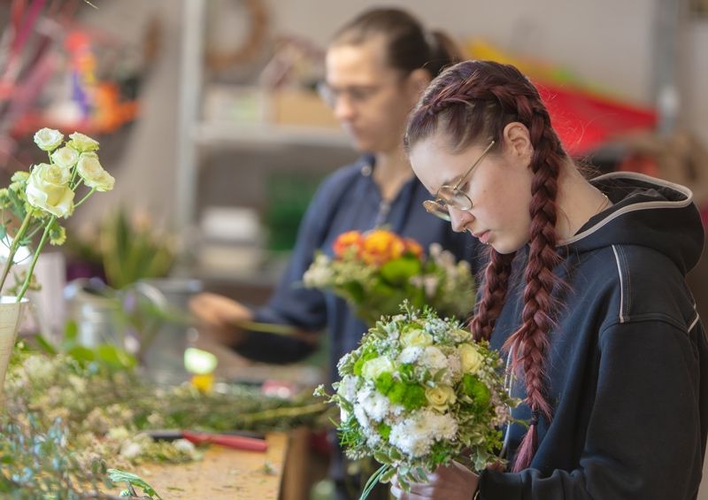Zwei Frauen sind in einem Raum mit Blumen, eine arrangiert einen Strauß, während die andere ihre Arbeit betrachtet.