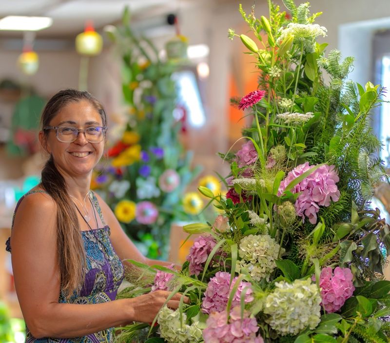 Eine Frau in einem Blumenkleid arrangiert eine große Blumenarrangement mit rosa und weißen Blumen in einem gut beleuchteten Raum.