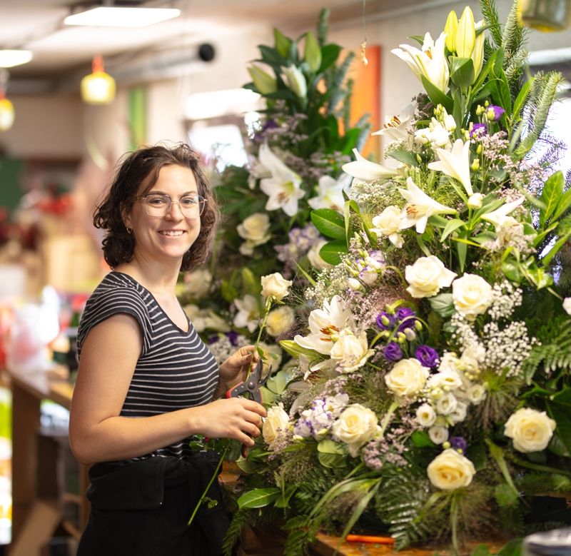 Eine Frau in einem Blumenladen, lächelnd und eine Schere haltend, arbeitet an einer großen Blumenarrangement.