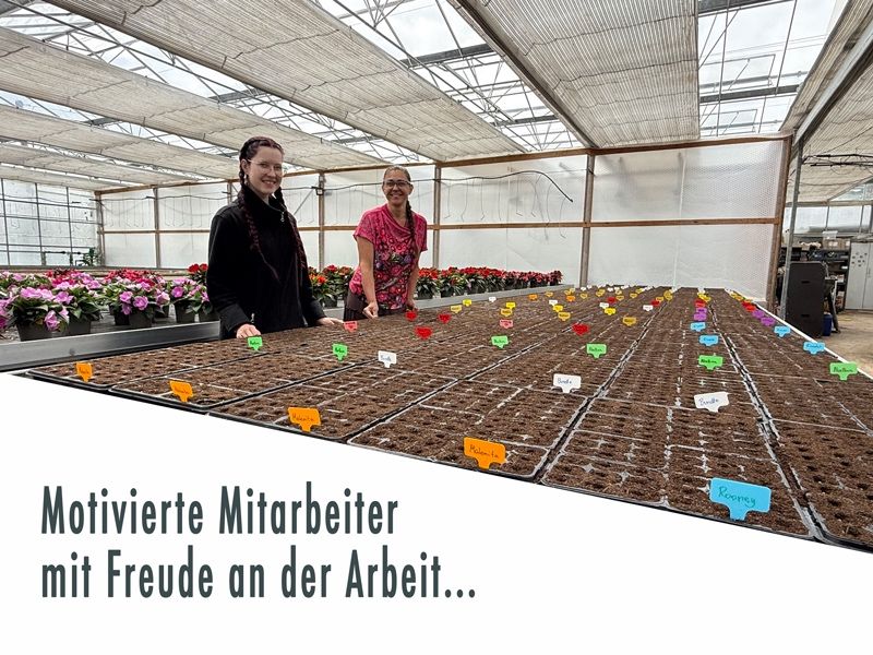 Zwei Frauen stehen lächelnd in einem Gewächshaus mit Reihen von Pflanzen. Auf den Pflanzen befinden sich Namensschilder. Das Gewächshaus hat Metallrahmen und eine weiße Decke.