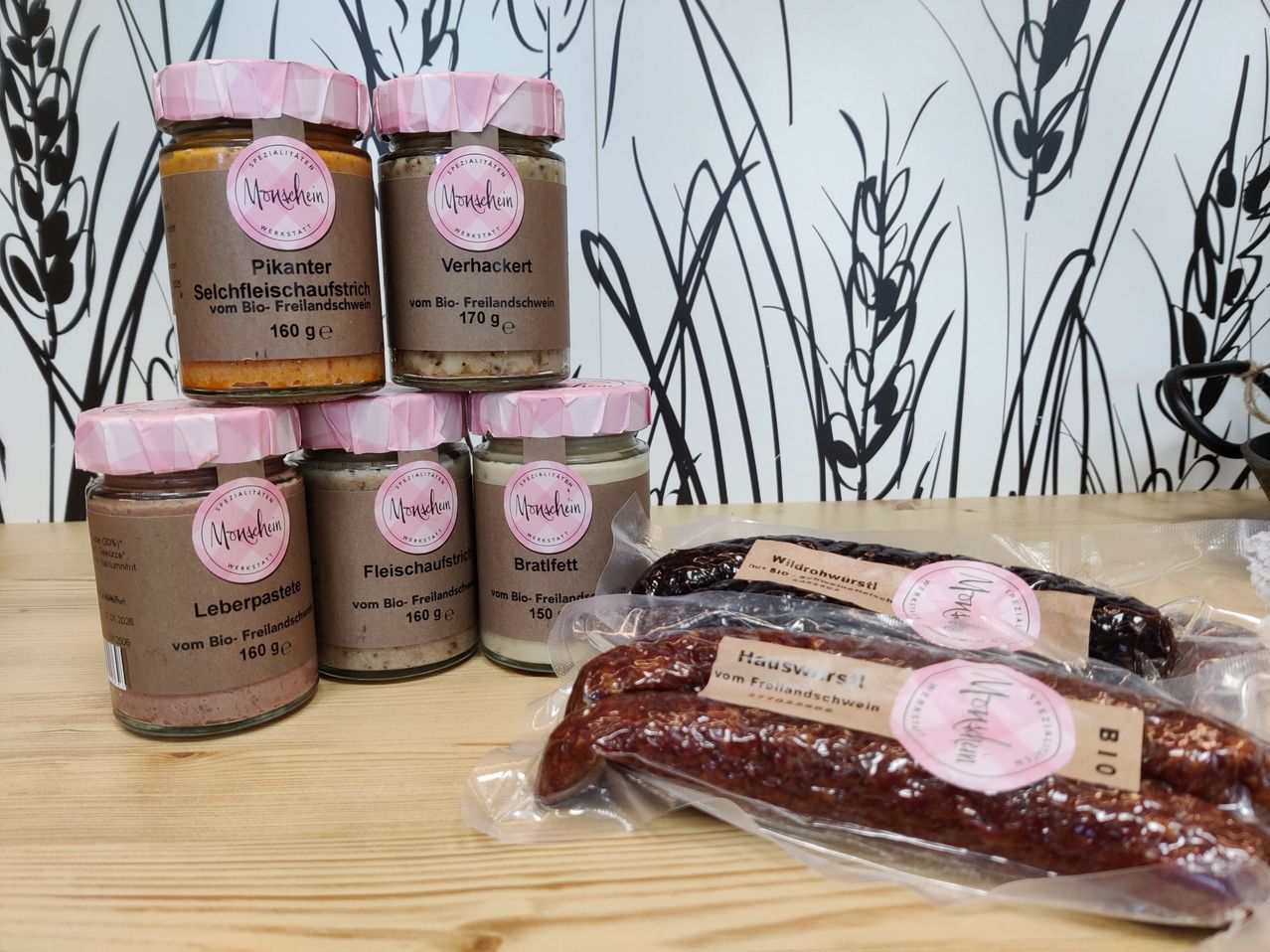 Mehrere Gläser mit verschiedenen Fleischprodukten sind auf einem Holztisch gestapelt, darunter Leberpastete, Verhackert und Bratfett. Darunter befinden sich Packungen mit Wildrohwurst und Hauswurst. Alle Artikel stammen von Bio-Freilandschwein.