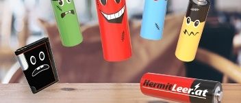 Fünf Batterien in verschiedenen Farben und Größen schweben auf einem Holztisch. Jede Batterie hat ein einzigartiges Gesicht darauf gezeichnet. Die rote Batterie hat ein lächelndes Gesicht. Die blaue Batterie hat ein überraschtes Gesicht. Die grüne Batterie hat ein trauriges Gesicht. Die gelbe Batterie hat ein schockiertes Gesicht. Die schwarze Batterie hat ein wütendes Gesicht.