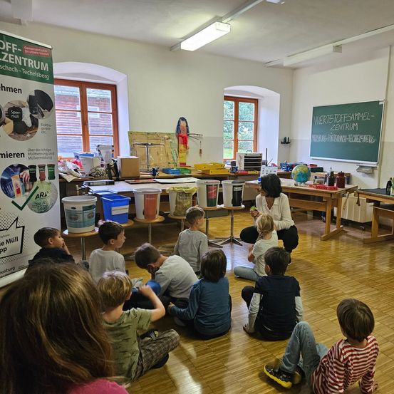 In einem Klassenzimmer sitzt eine Lehrerin auf dem Boden, umgeben von aufmerksam zuhörenden Kindern. Der Raum ist gut beleuchtet und enthält Schreibtische, Aufbewahrungsboxen und eine Tafel mit deutschem Text.