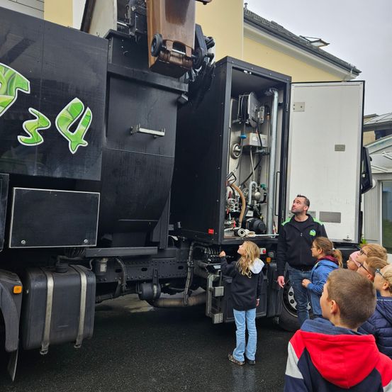 Ein Mann und Kinder betrachten einen schwarzen Lkw mit der Nummer 354 an der Seite. Der Lkw hat eine offene Tür, die seine Maschinerie zeigt.