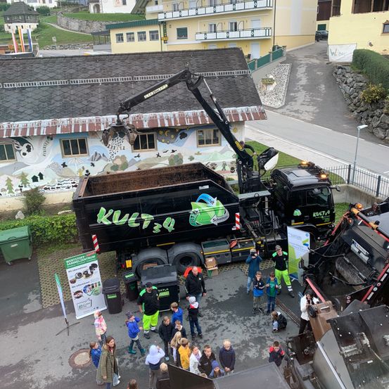 Ein großer Lkw mit der Aufschrift kult34 wird von einem Kran bedient. Viele Menschen beobachten ihn. Einige Mülleimer stehen auf der linken Seite. Hinter dem Lkw befinden sich einige Gebäude. Einige Straßenlaternen stehen auf der rechten Seite.