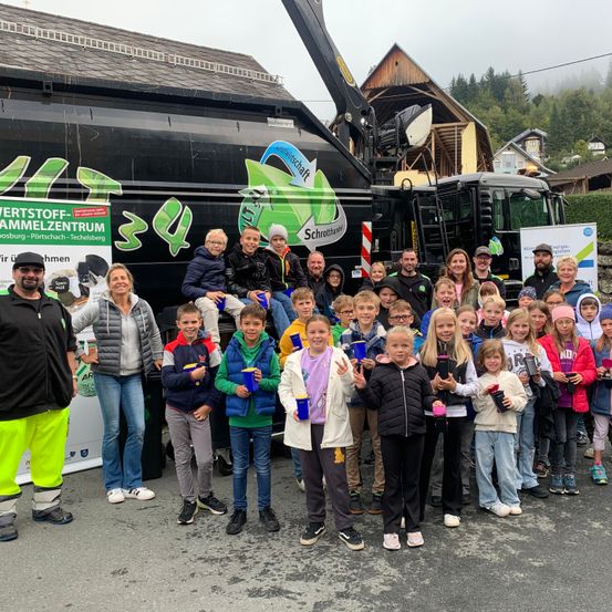 Eine Gruppe von Menschen, darunter Kinder, steht vor einem Lastwagen mit Recycling-Logo. Einige halten Becher in der Hand, und es gibt Erwachsene und Kinder in verschiedenen Outfits. Im Hintergrund sind Häuser und Bäume zu sehen.