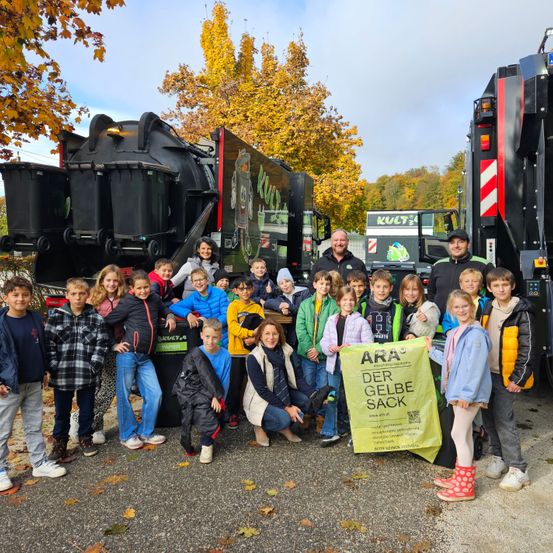 Eine Gruppe von Kindern und Erwachsenen steht vor Müllwagen, einer hält ein gelbes Banner mit der Aufschrift 'ARA DER GELBE SACK'. Die Kinder tragen Jacken, einige mit Mützen, und Turnschuhe. Die Trucks haben schwarze Behälter auf ihnen.