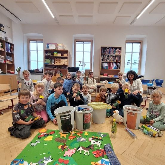 Kinder versammeln sich in einem Klassenzimmer mit einem Bauernhoftier-Teppich, halten grüne Behälter und verschiedene Gegenstände. Zwei Frauen und eine Ente sind anwesend, mit Regalen voller Bücher und Gegenständen im Hintergrund.