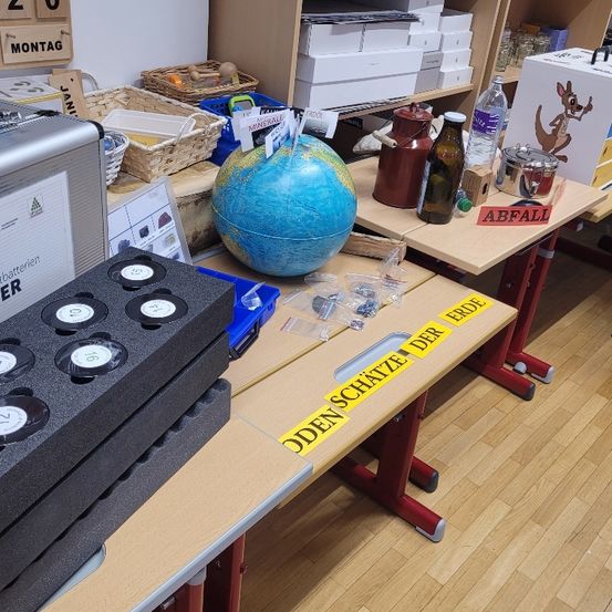 Ein Raum zeigt einen Schreibtisch mit einer Globus, Flaschen und einem Schild mit der Aufschrift 'Den Schatze Der Erde'. Ein anderer Tisch hat Aufbewahrungsboxen und Körbe. Die Etiketten 'Abfall' und 'Montag' sind sichtbar. Ein Kasten mit einem Känguru ist rechts platziert.