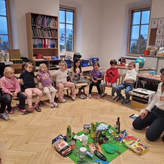 Ein Klassenzimmer mit Kindern, die in einem Kreis um einen Tisch sitzen, der mit verschiedenen Gegenständen gefüllt ist, während eine Frau auf dem Boden kniet.