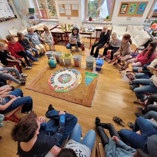 Ein Klassenzimmer mit Kindern, die in einem Kreis um ein Brettspiel auf dem Boden sitzen. Eine Frau sitzt in der Nähe.