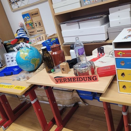 Ein Klassenzimmer mit zwei Tischen mit verschiedenen Gegenständen darauf. Ein Globus, eine Flasche und eine rote Box mit dem Wort 'Vermeidung'. Ein Regal mit Kisten und einem Kalender.