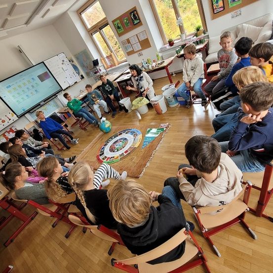 Eine Gruppe von Kindern in einem Klassenzimmer sitzt um einen Tisch mit einem Globus herum und spielt ein Spiel auf dem Boden.