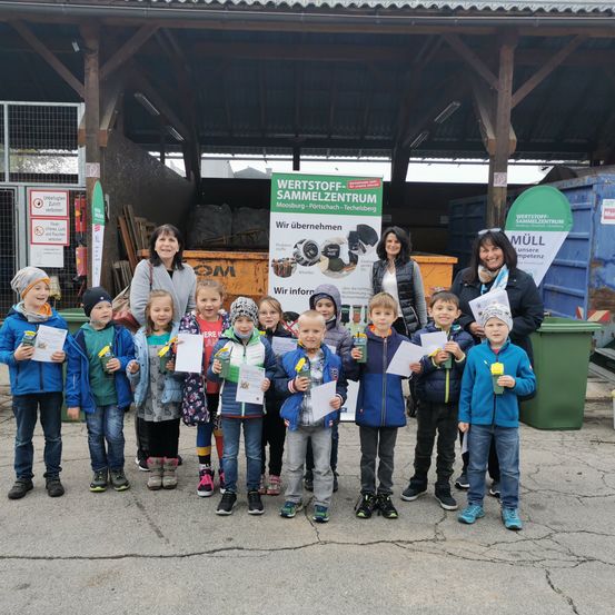 Eine Gruppe von Kindern und Erwachsenen steht vor einem Wertstoff Sammelzentrum. Die Kinder halten Zertifikate. Zwei Frauen stehen hinter ihnen.