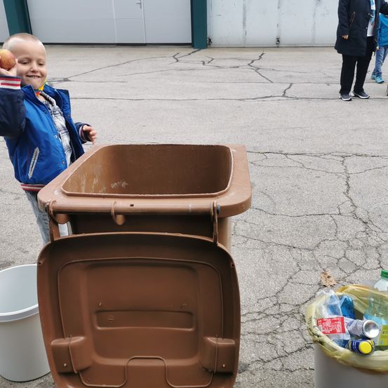 Ein junger Junge mit einem Apfel in der Hand steht neben einem offenen Mülleimer im Freien. Dahinter laufen zwei Personen auf einer rissigen Straße.