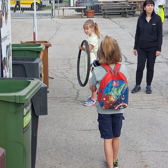 Zwei Kinder und eine Frau gehen in einem Park. Ein Kind hält einen Reifen und ein anderes trägt einen Rucksack mit Superhelden. Mülleimer und Bänke befinden sich hinter ihnen.