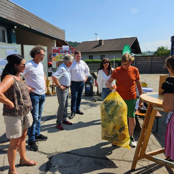 Eine Gruppe von Menschen steht vor einem Gebäude und hält einen gelben Sack mit der Aufschrift 'ARA DER GELB SACK'. Einige Personen betrachten einen Holztisch mit Gegenständen. Ein Gebäude mit Schornstein befindet sich hinter ihnen.