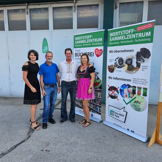 Vier Personen stehen vor einem Gebäude mit Bannern. Die beiden Banner auf der rechten Seite informieren über Abfallmanagement und Recycling.