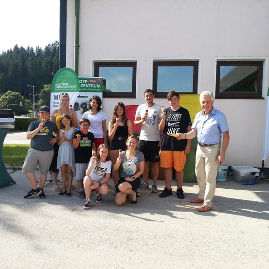 Eine Gruppe von Menschen steht vor einem Gebäude. Sie halten Eiscreme-Eis. Hinter ihnen ist ein Banner. Im Hintergrund befindet sich ein Gebäude mit drei Fenstern. Bäume und Büsche sind in der Ferne.