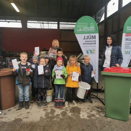 Eine Gruppe von Kindern, Erwachsenen und einer Frau stehen in einem Recycling-Bereich, halten Papiere und Tassen in der Hand, mit zwei grünen Mülleimern und einem Banner hinter ihnen.