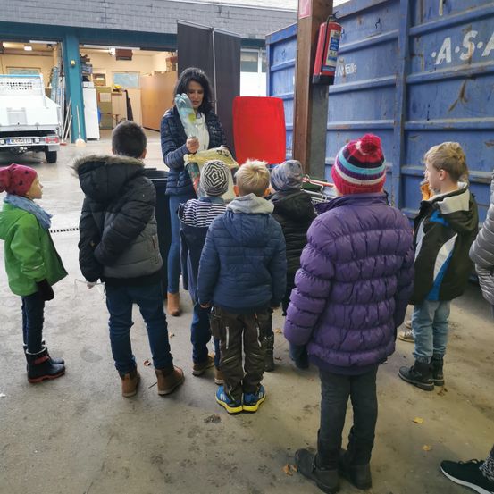 Eine Frau hält einen Beutel mit Lebensmitteln, umgeben von Kindern in Winterkleidung. Sie stehen in einer Garage mit einem Feuerlöscher an der Wand.