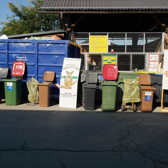 Verschieden farbige Mülltonnen sind vor einem Gebäude aufgereiht. In der Mitte steht ein Schild mit einem Känguru.