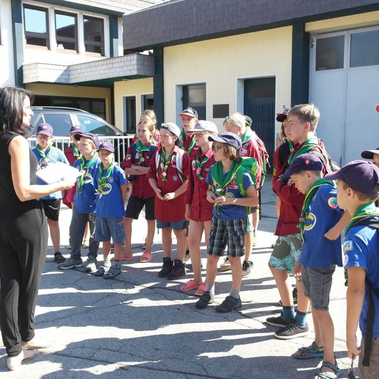 Eine Gruppe junger Pfadfinder, gekleidet in roten und blauen Uniformen, versammelt sich zu einer Versammlung, wobei eine Frau vorne aus einem Papier liest. Sie stehen vor einem Gebäude.
