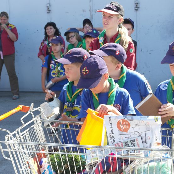 Eine Gruppe von Kindern in blauen Uniformen und Mützen ist in einem Einkaufswagen mit Gegenständen. Sie sind von Erwachsenen im Hintergrund umgeben. Ein Kind hält ein Buch.