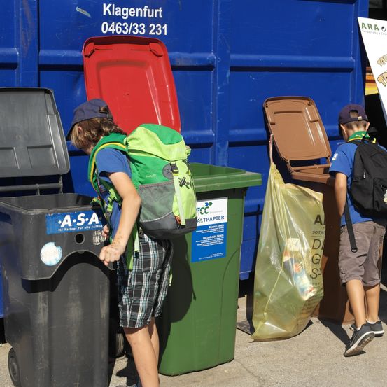 Zwei Kinder stehen neben Recyclingbehältern, eines mit einem Rucksack, der Müll in einen Behälter wirft. Die Behälter sind grün und braun, wobei der blaue Behälter mit 'Klagenfurt 0463/33 231' beschriftet ist.