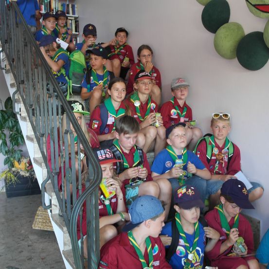 Eine Gruppe junger Pfadfinder in roten Uniformen und Mützen sitzt auf einer Treppe, einige essen Snacks, und sie stehen vor einer Wand mit hängenden grünen Dekorationen.