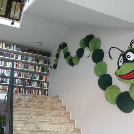 Eine Treppe führt zu einem Balkon mit Geländer, und ein grüner Raupenplüsch schmückt die Wand. Ein Regal mit Büchern steht links.