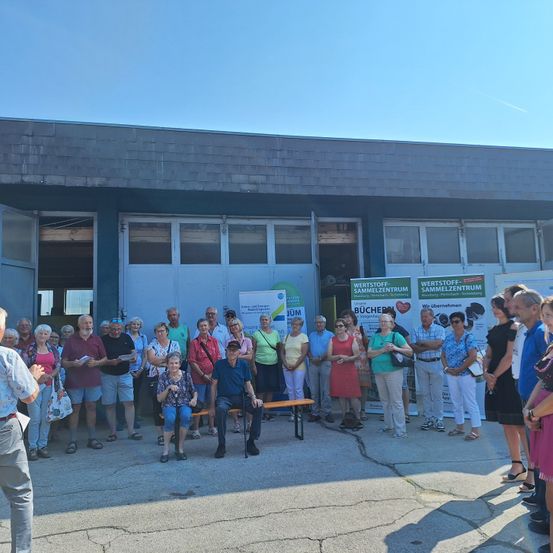 Eine Gruppe von Menschen, darunter Männer und Frauen, steht und sitzt vor einem Gebäude, möglicherweise für eine Veranstaltung. Sie tragen Freizeitkleidung. Das Gebäude hat Glasfenster und eine Tür.