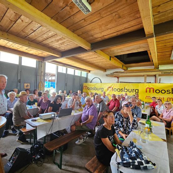 Eine Gruppe älterer Menschen sitzt in einem Saal an Tischen und schaut sich etwas auf einem Laptop an. Eine Fahne mit dem Wort Moosburg hängt an der Wand.