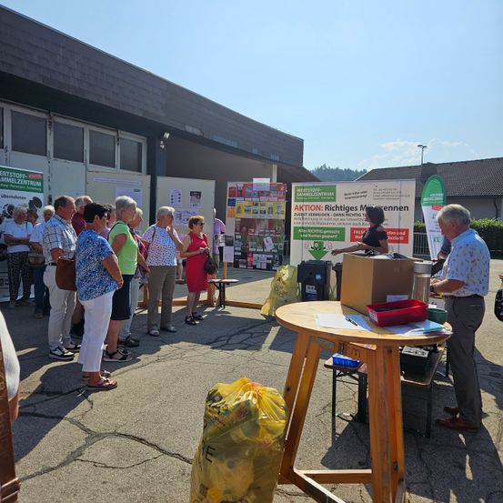 Eine Gruppe von Menschen versammelt sich vor einem Gebäude mit Schildern, eine Person steht bei einem Tisch mit einem Karton und Müllsäcken.