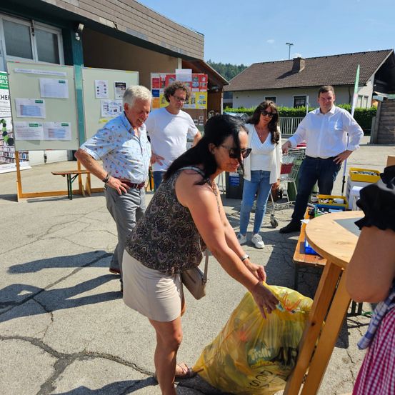 Eine Gruppe von Menschen steht vor einem Gebäude. Eine Frau hält einen gelben Beutel. Ein Mann in einem weißen Hemd steht neben ihr. Ein anderer Mann hält einen Einkaufswagen. Sie stehen vor einem Tisch mit einem gelben Beutel darauf.